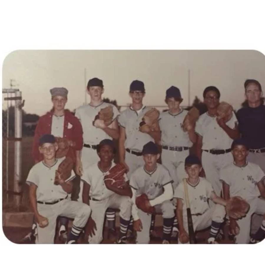 A vintage photo of a youth baseball team posing on a field. Eight boys in uniforms, one boy in a red jacket, and an adult are grouped together, smiling and holding gloves.