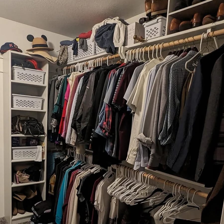Walk-in closet filled with hanging clothes, organized by color and type. Shelves hold white baskets, hats, shoes, and bags. A hat with mouse ears sits on the top left shelf. The closet is neatly arranged and well-lit.