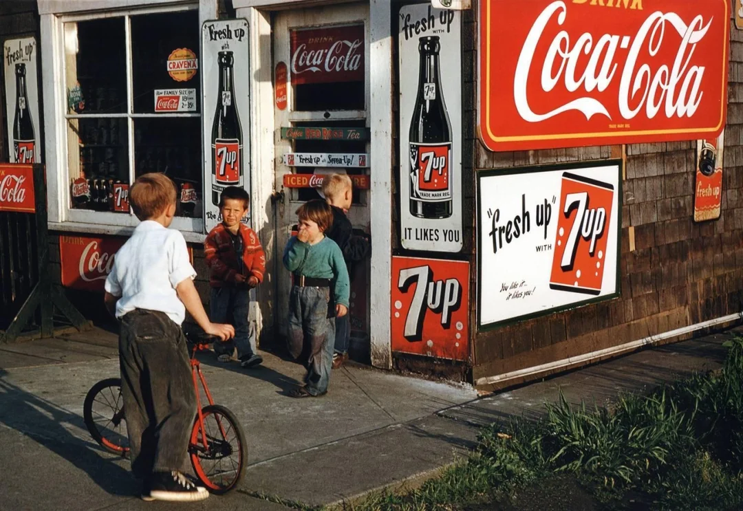 Four children stand and chat outside a small store covered in Coca-Cola and 7 Up signs. One child holds a red scooter, and the store’s door and window are partially open. The scene appears to be from the mid-20th century.