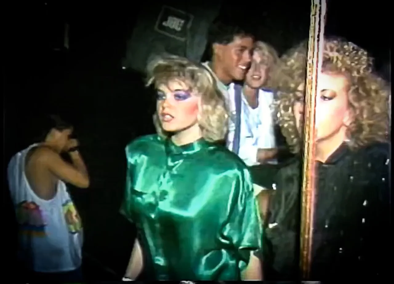A woman in a shiny green blouse with bold makeup stands in front of a mirror. Other people, also dressed in vintage 1980s fashion, smile and talk in the background. The scene has a retro nightlife vibe.