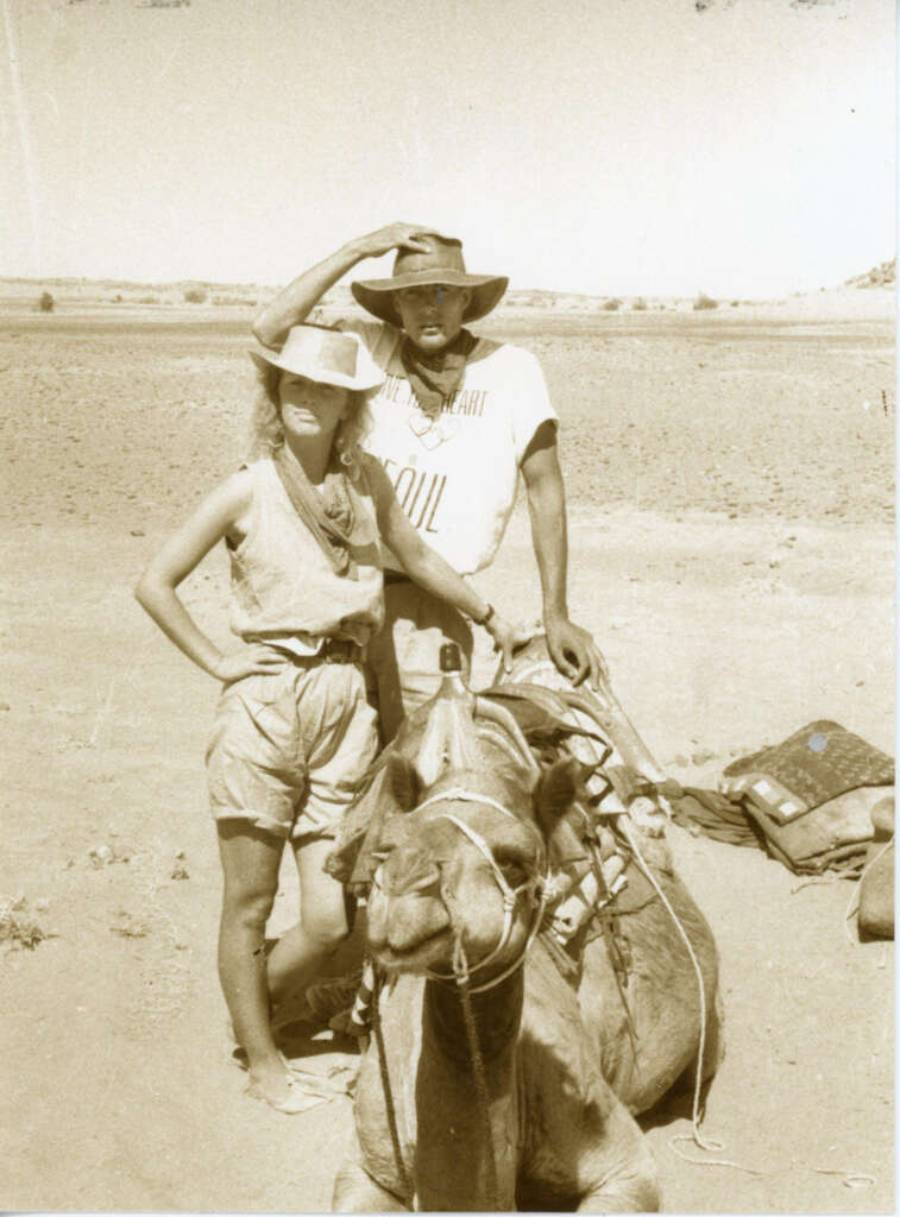 Two people in hats stand in a desert beside a sitting camel. One person rests their hand on the other’s hat. There is luggage on the ground nearby. The photo is sepia-toned, giving it a vintage look.