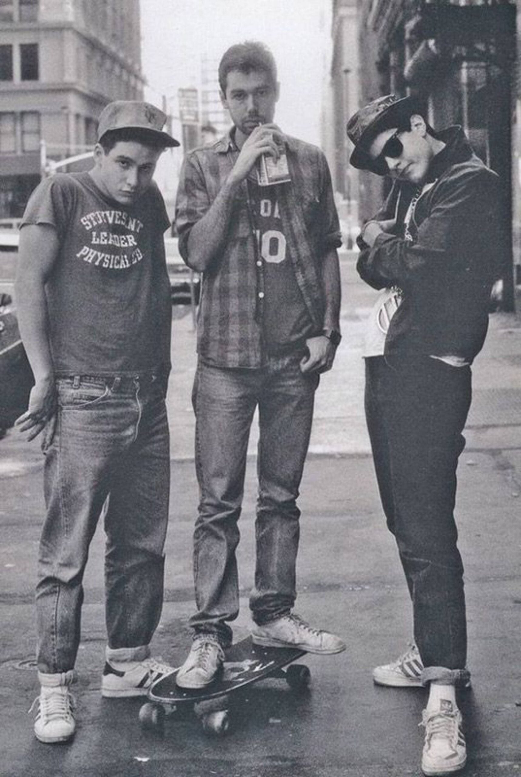 Three young men pose casually on a city street; one stands on a skateboard, another drinks from a can, and the third crosses his arms and wears sunglasses and a hat. All wear jeans and sneakers.