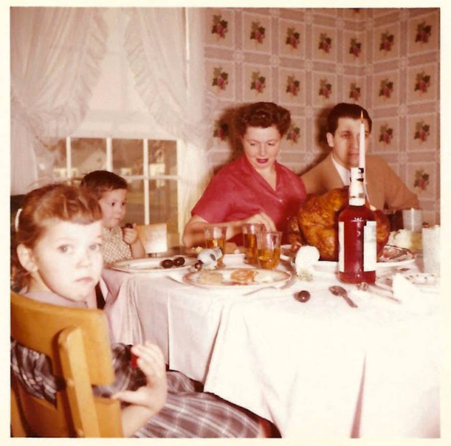 A family of four sits at a dining table with a large roasted turkey, drinks, and food. The mother serves food, while the father and two young children sit nearby in a vintage-style kitchen.