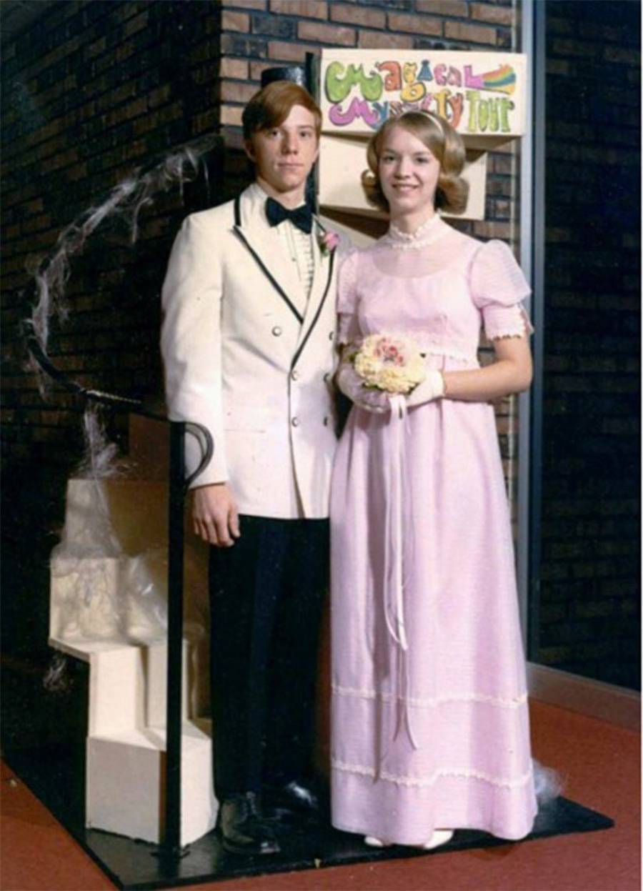 A young couple poses indoors; the boy wears a white tuxedo and the girl wears a pink dress, holding flowers. A “Magical Mystery Tour” sign is behind them on a brick wall.
