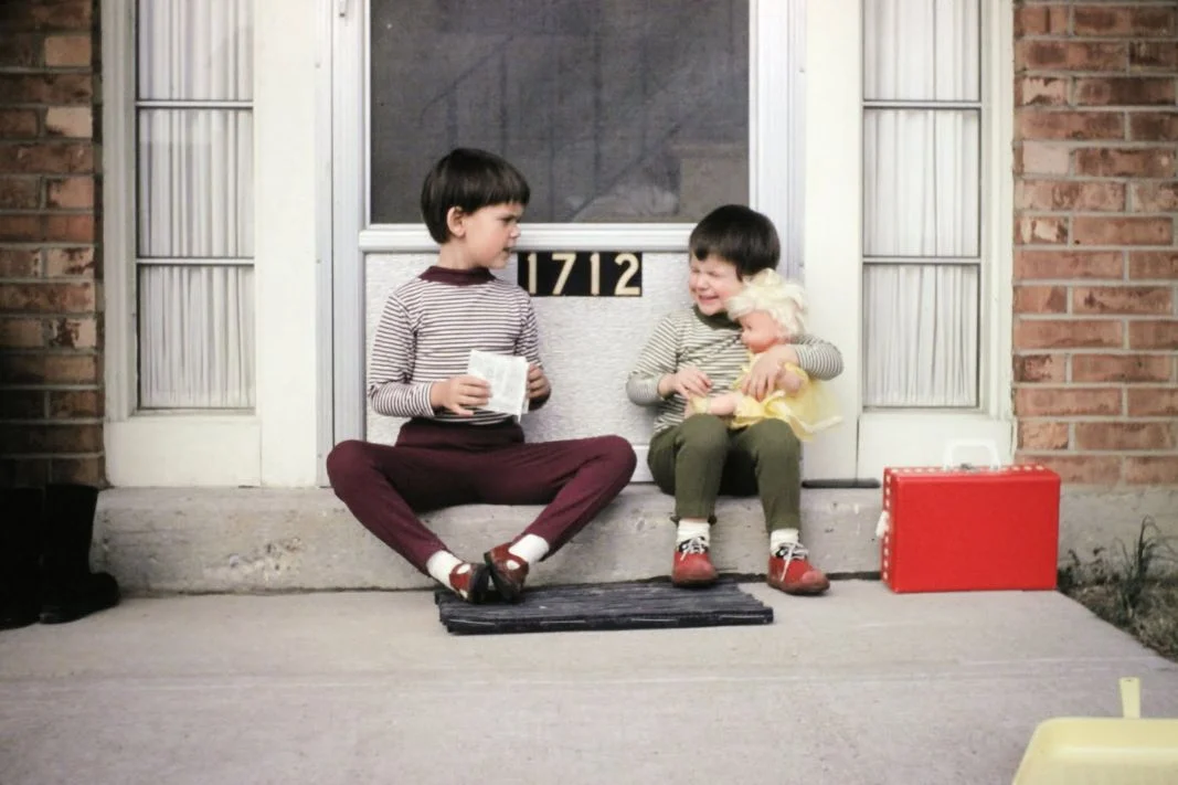 Two children sit on a doorstep in front of a door numbered 1712. One holds papers, the other holds a doll. Both wear striped shirts and red shoes. A red box and yellow toy are nearby.