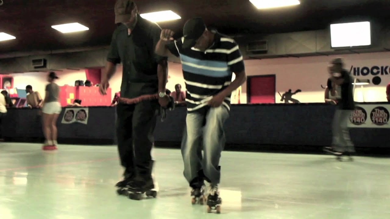 Two men roller skate indoors, one in a striped shirt appearing to dance, while others skate in the background under fluorescent lights.
