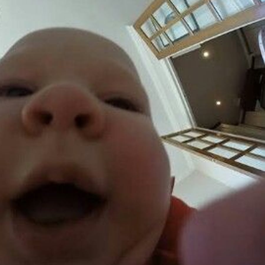 A baby’s face close to the camera, mouth open, with a ceiling and open doors visible in the background.