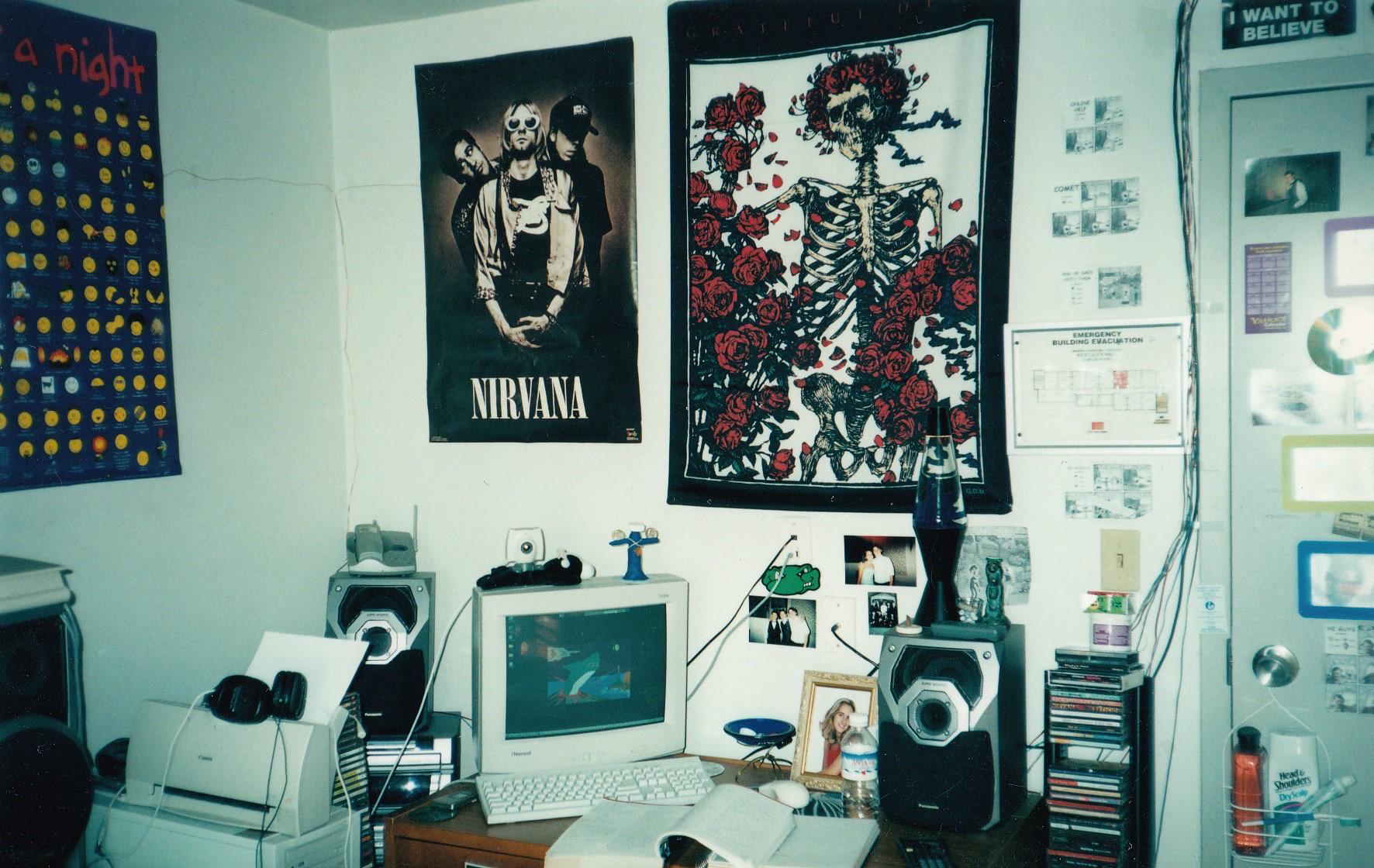 A cluttered desk with an old computer, speakers, CDs, and headphones. Nirvana and skeleton tapestry posters hang on the wall. Papers, photos, and various tech accessories fill the small, busy room.