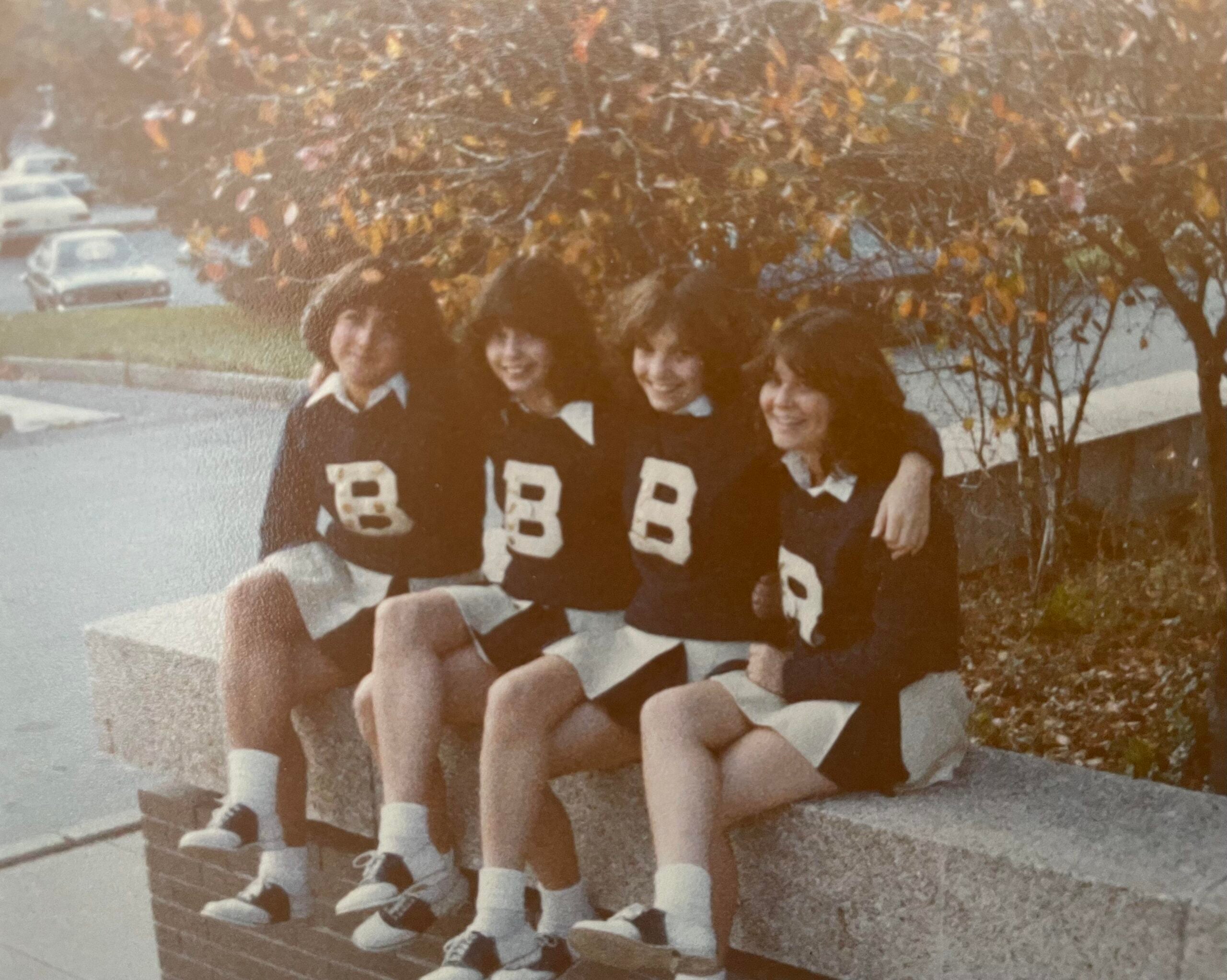 Four girls in matching cheerleader uniforms with letter “B” on sweaters sit on a low wall outside, smiling with arms around each other. Trees and parked cars are visible in the background.