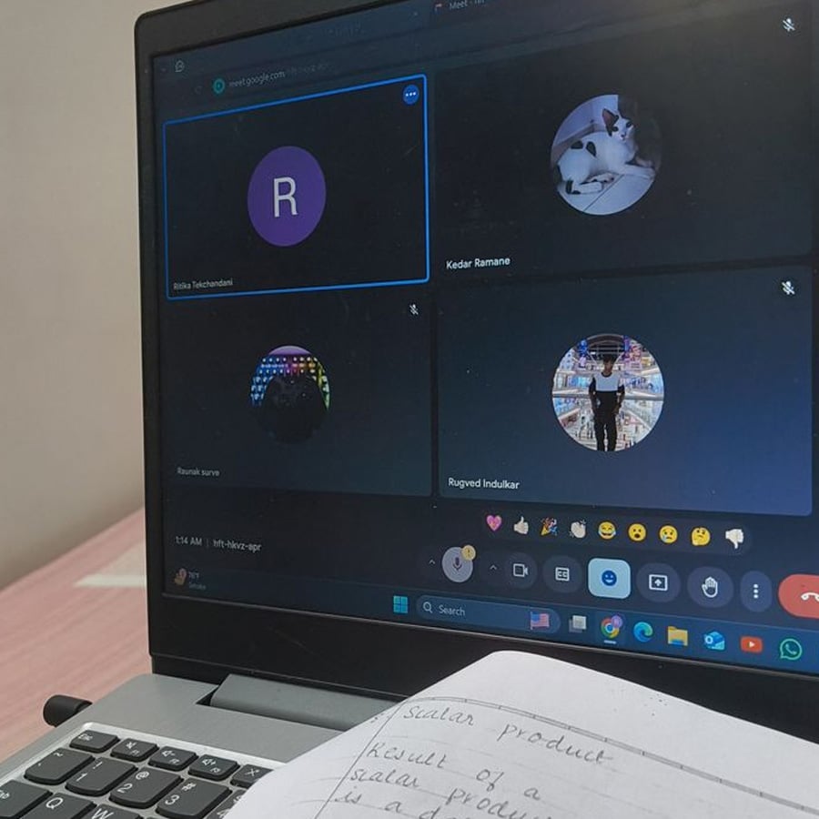A laptop displays a virtual meeting with four participants, some using avatars. In front of the laptop, a notebook with handwritten notes is visible on a pink desk.