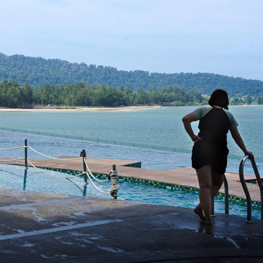 A person stands at the edge of a pool, holding the railing, looking out toward a large body of water and distant forested hills under a clear sky.