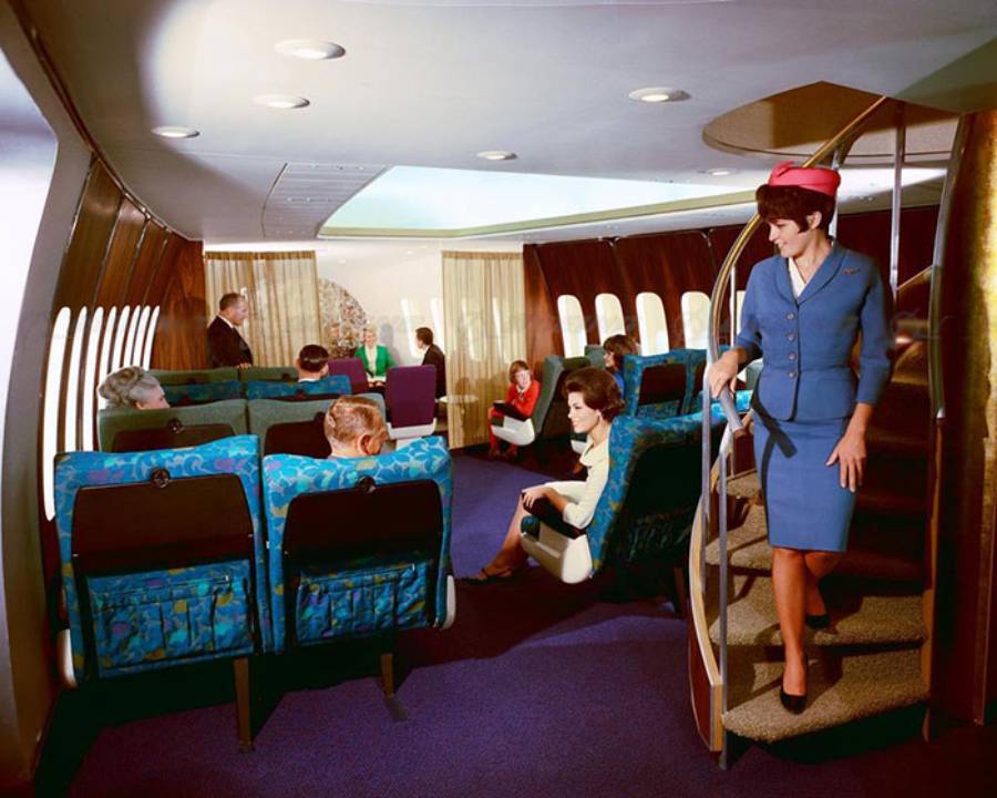 Vintage airplane interior with passengers seated in spacious, colorful seats. A flight attendant in a blue uniform and hat descends a spiral staircase. Large windows and relaxed atmosphere suggest 1960s or 1970s air travel luxury.