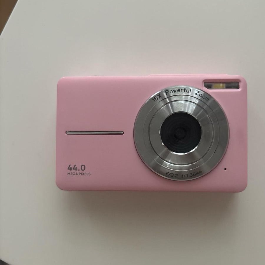 A pink digital camera with a large lens, labeled “16X Powerful Zoom” and “44.0 Mega Pixels,” sits on a white surface. The flash and shutter button are visible.
