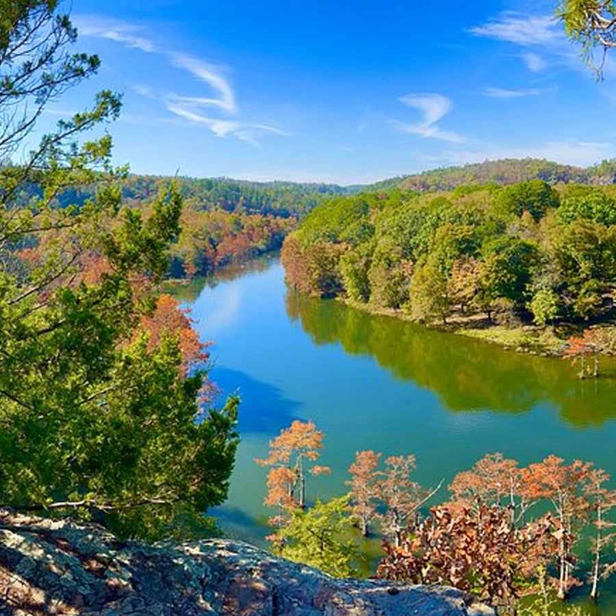 A scenic river winds through a lush, green forest under a bright blue sky with wispy clouds, viewed from a rocky, elevated vantage point.
