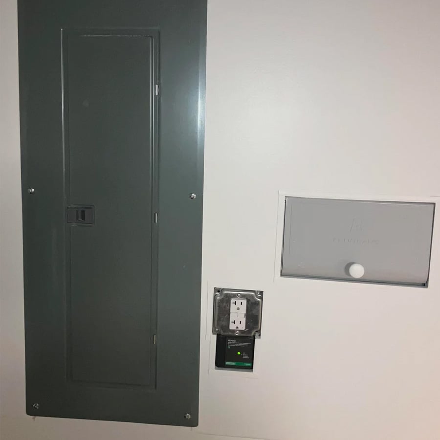 A gray electrical panel, a small gray utility door, and an electrical outlet with a green light are mounted on a white wall.