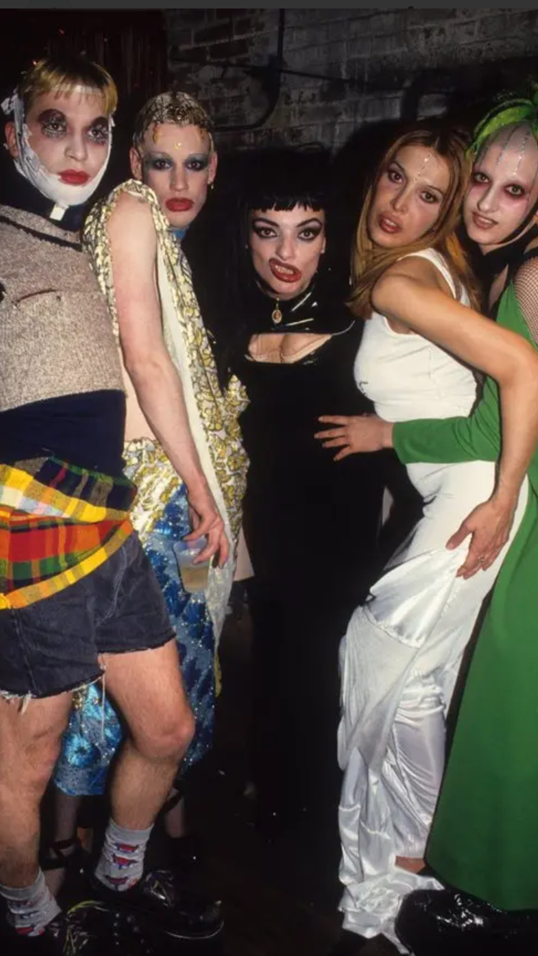 Five people in dramatic, colorful costumes and makeup pose together at a party. Their bold outfits include sequins, fishnets, and eclectic fabrics, and they stand closely, making playful expressions for the camera.