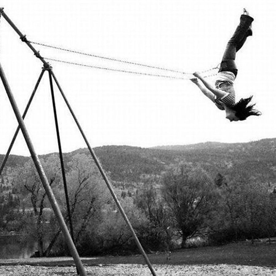 A person swings high on a swing set, upside down in midair, holding onto the chains. Trees, hills, and a lake are visible in the background.