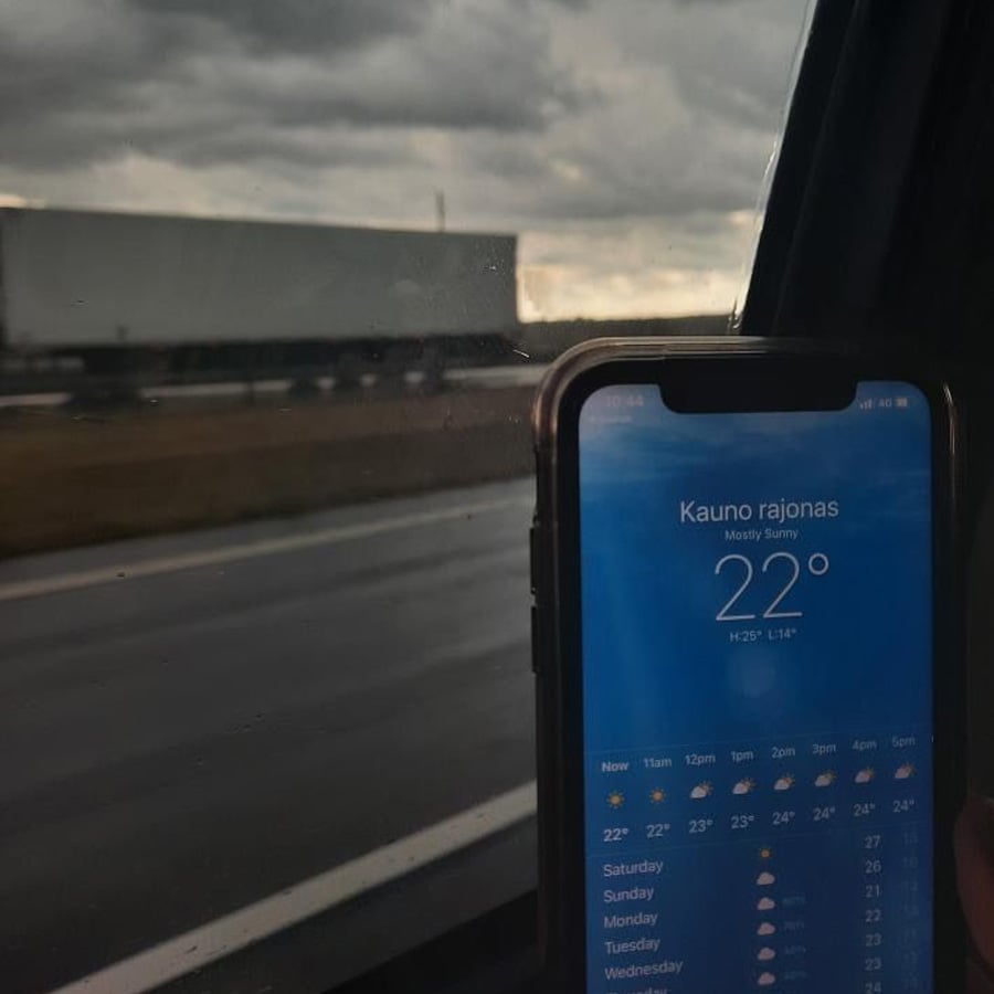 A smartphone displays a weather app showing 22°C and “Kauno rajonas, Mostly Sunny” against a rainy highway scene viewed through a car window.