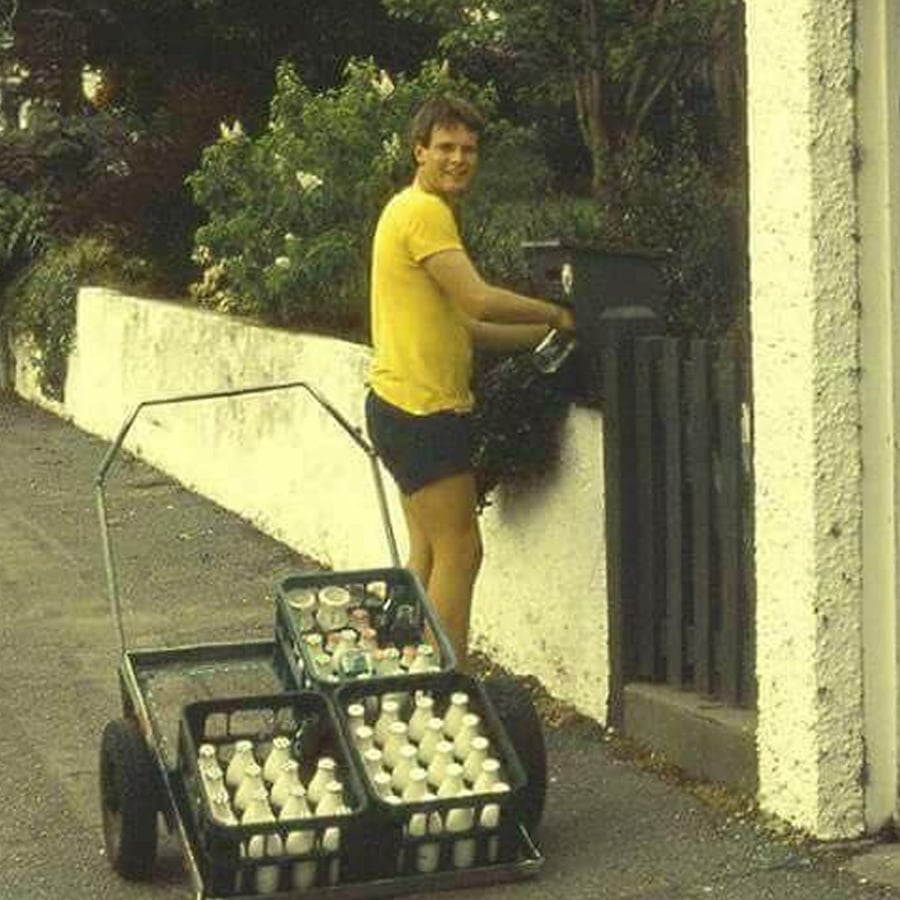 A man in a yellow shirt and shorts delivers milk bottles with a cart, stopping at a gate to place bottles in a box. The scene appears to be from a past decade based on the outfit and milk bottles.