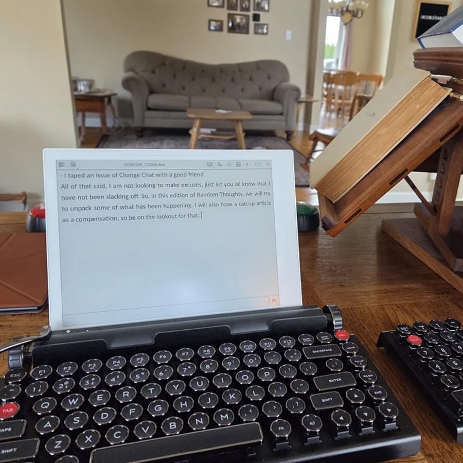 A tablet with a keyboard shows a text document, set on a desk with notebooks. In the background, a living room with a sofa, chairs, and a bookshelf can be seen.
