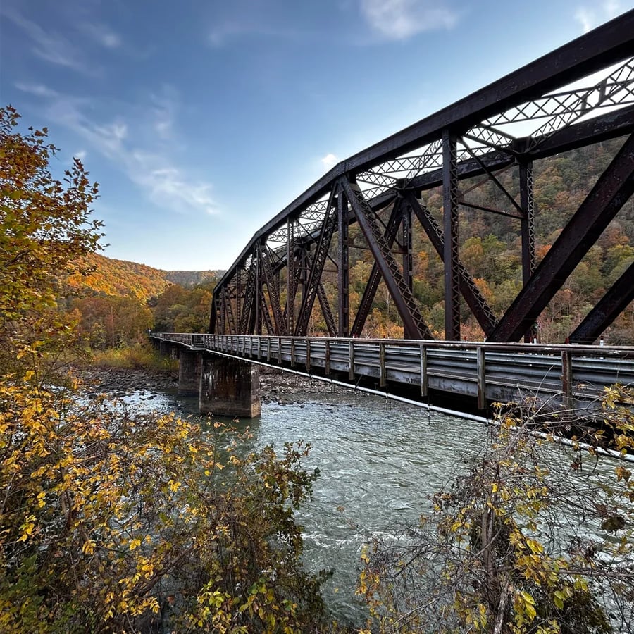A black steel truss bridge spans a river, surrounded by autumn trees. The sky is clear and blue, with forested hills visible in the background.