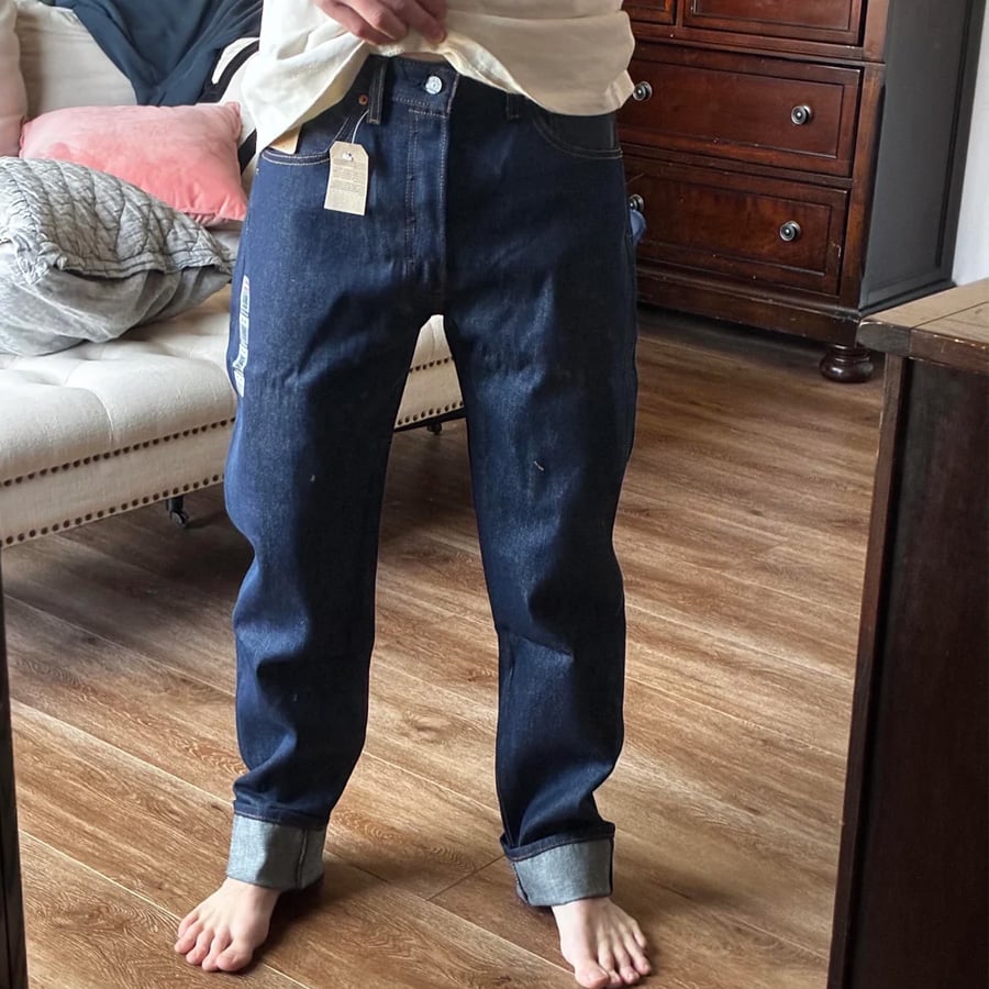 A person wearing dark blue cuffed jeans and a white shirt stands barefoot on a wooden floor in a bedroom, taking a mirror selfie.