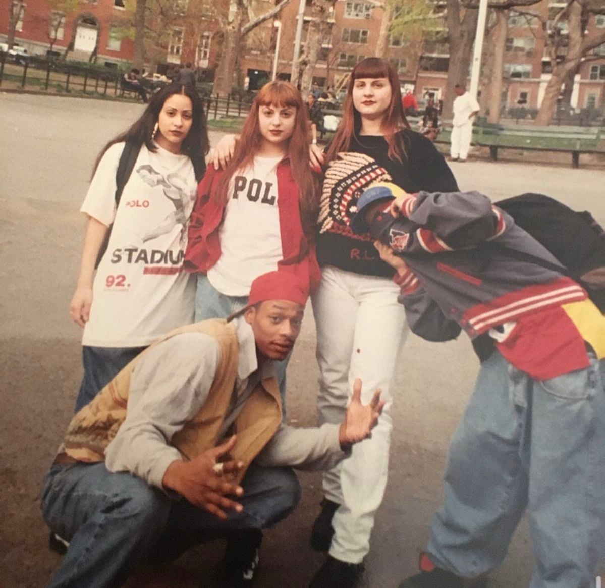 Five young people pose together in an outdoor park. Three stand in back, while two kneel in front. They wear casual ’90s clothing, including Polo shirts, jeans, and caps. Trees, a fence, and buildings are visible in the background.