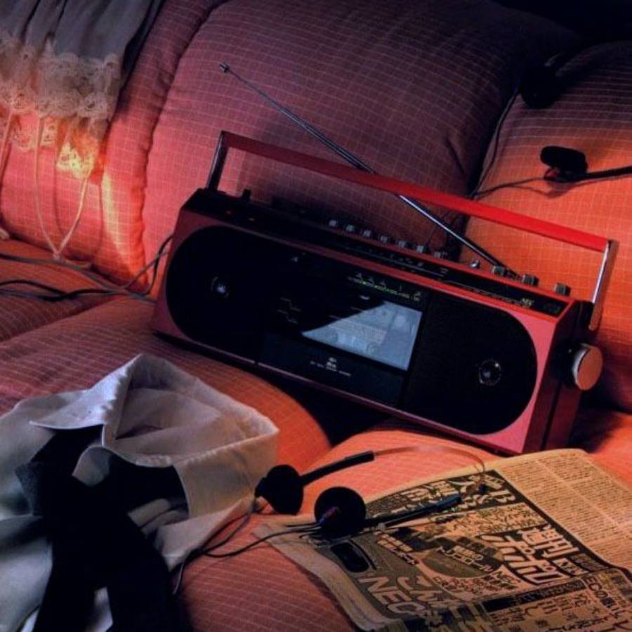 A red radio sits on a pink, quilted bed with a shirt, tie, headphones, and a newspaper scattered around it, creating a cozy, retro atmosphere.