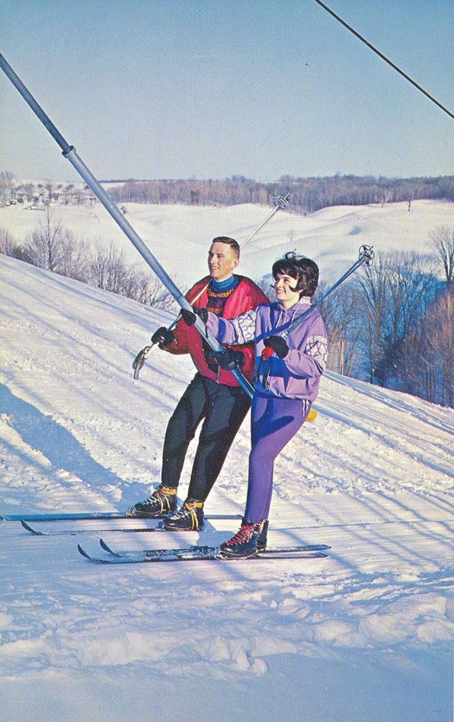 A man and woman in colorful ski outfits ride a ski lift together up a snowy hill, holding onto the lift pole, with trees and a clear sky in the background.