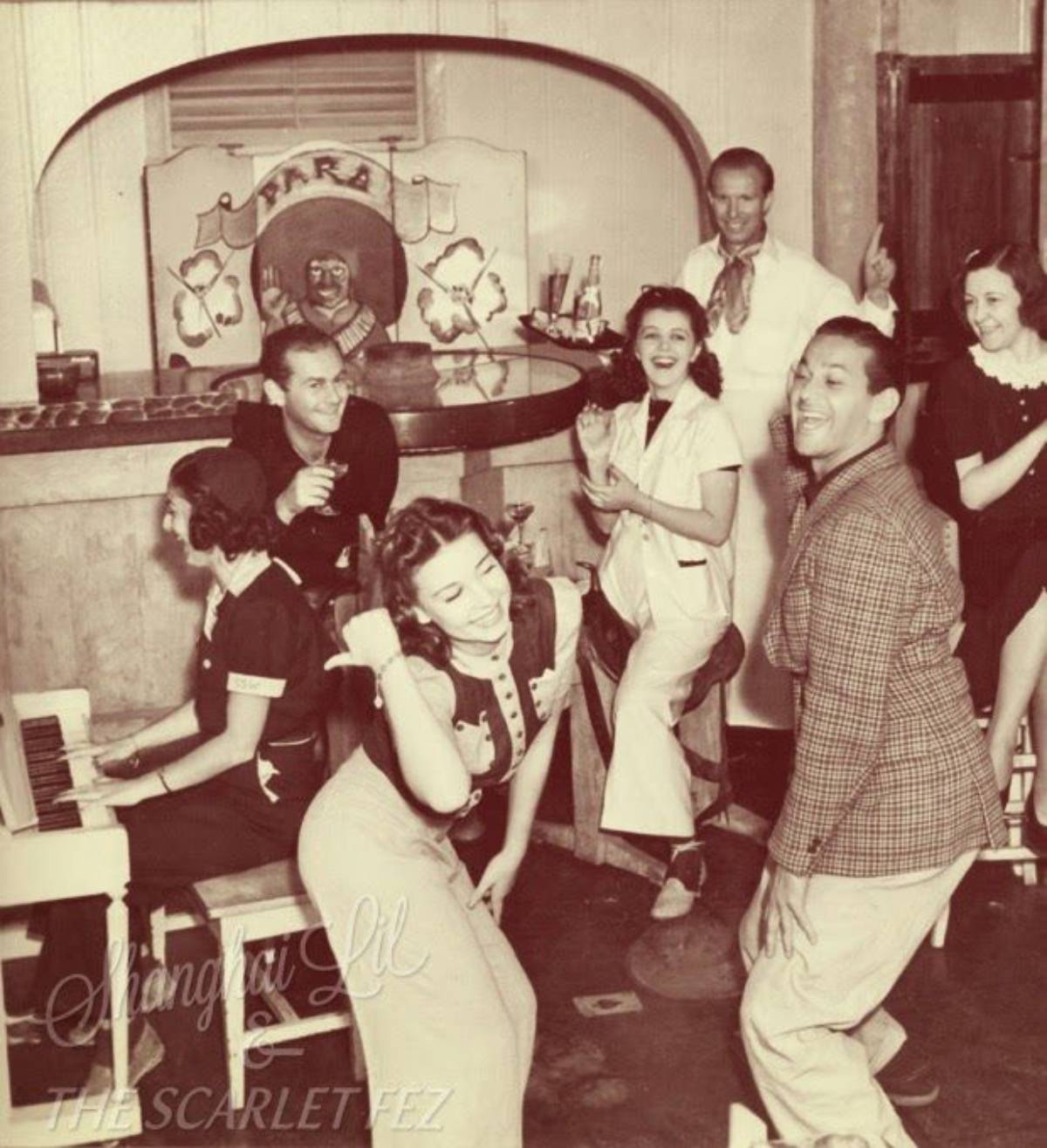 A vintage scene shows people dancing and singing around a piano in a lively bar, with a bartender in the background. Everyone appears joyful, enjoying the music and each other’s company.