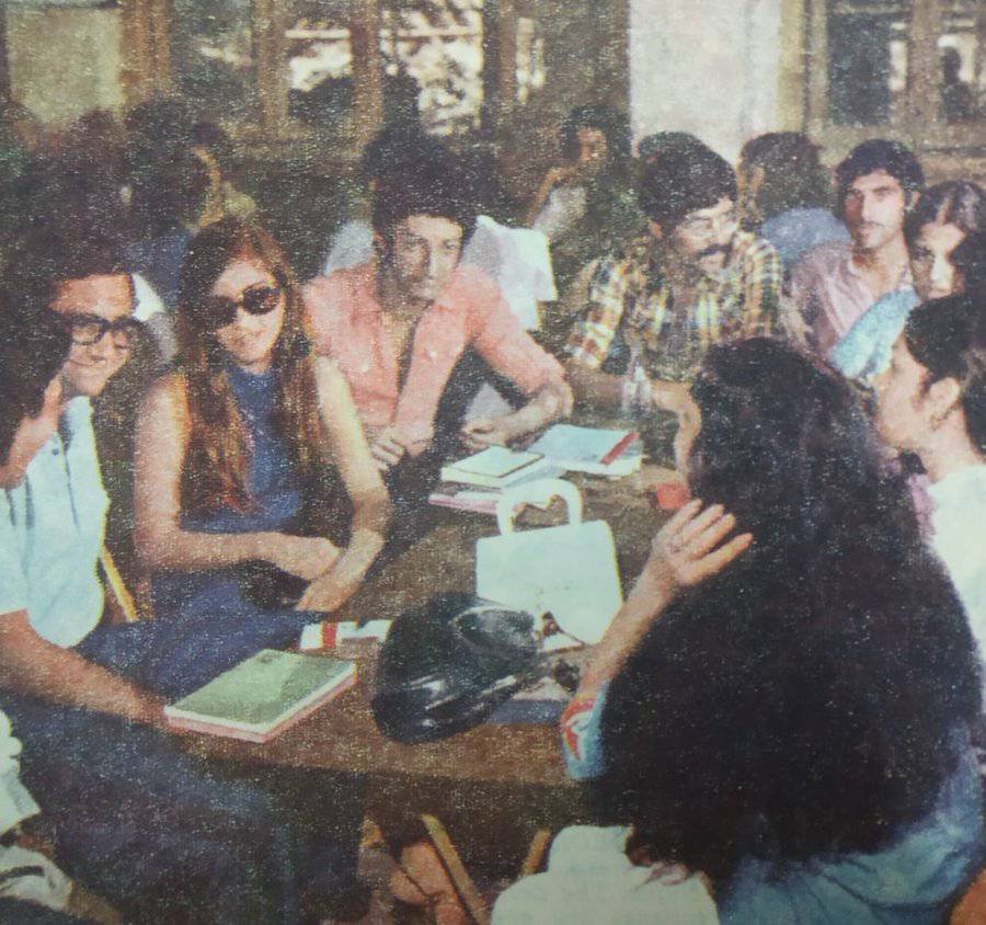 A group of young people sit around a round table, talking and reading books. Some wear glasses, one has sunglasses, and notebooks and bags are on the table. The setting appears to be indoors, possibly a library or café.