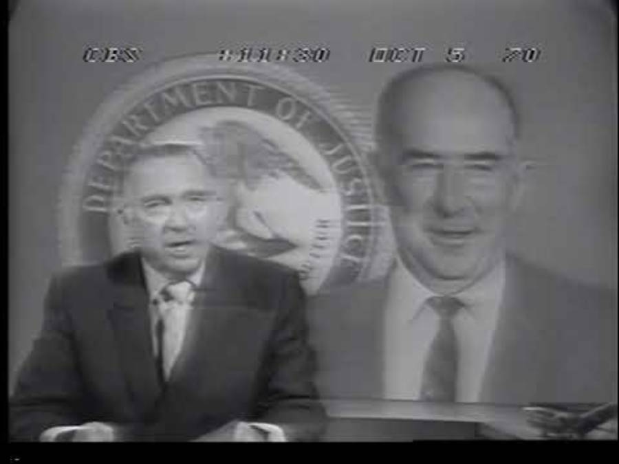 A news anchor speaks on TV, with a large Department of Justice seal and a man’s portrait in the background. The date “OCT 5 70” and “CBS” appear at the top of the black-and-white image.