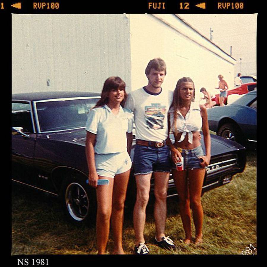 Three people in 1980s summer outfits stand in front of a dark classic car at an outdoor event. There’s a white building in the background and other people visible near more cars.