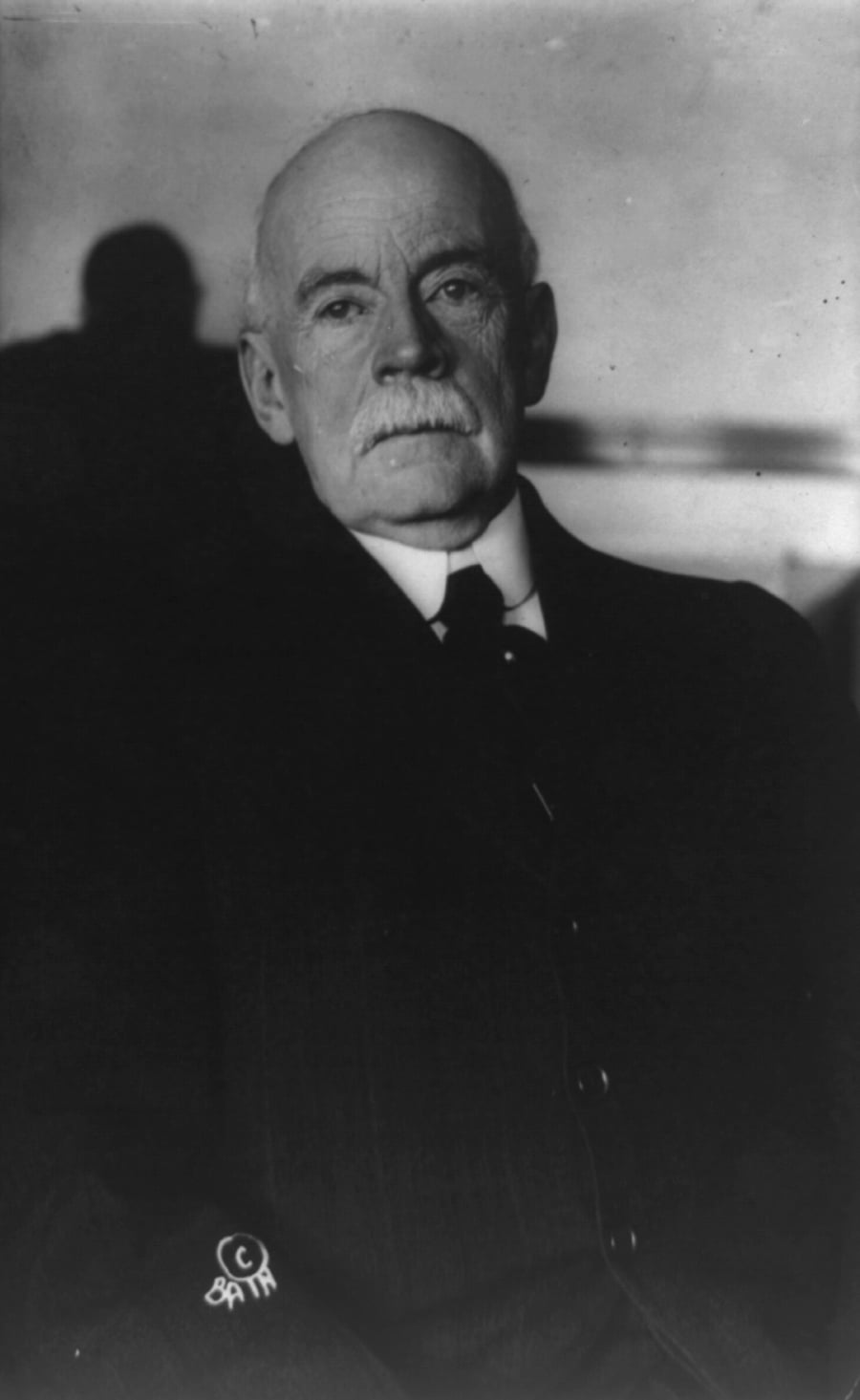 An older man with a mustache and bald head, wearing a dark suit and tie, sits facing the camera against a plain background.
