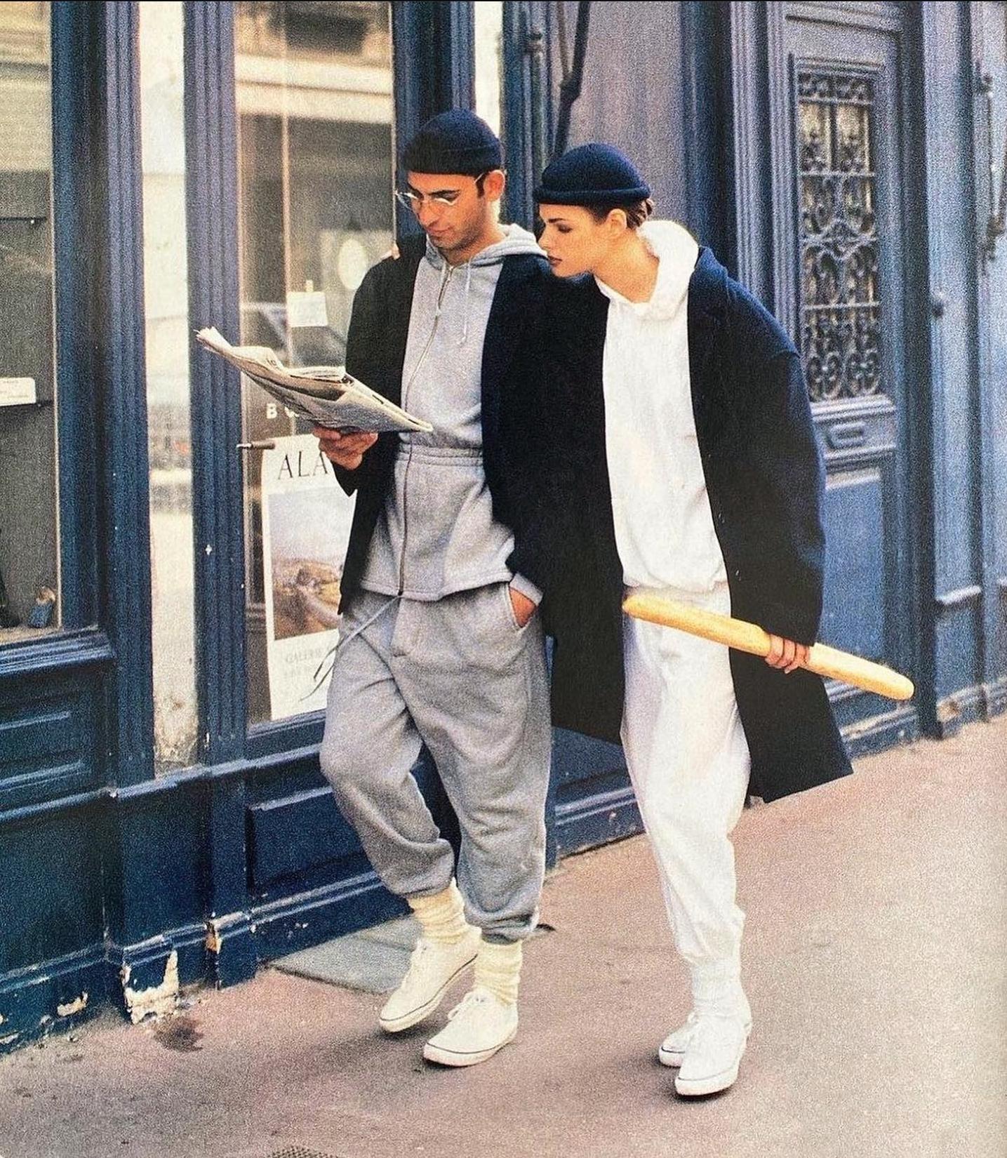 Two people in casual tracksuits and beanies walk on a city sidewalk; one reads a newspaper while the other holds a baguette. They pass in front of a dark blue storefront.