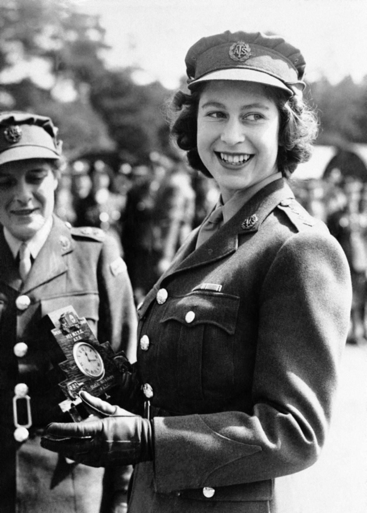 A young woman in a military uniform and cap smiles while holding a small framed clock. Another woman in uniform stands beside her. There are blurred people and trees in the background.