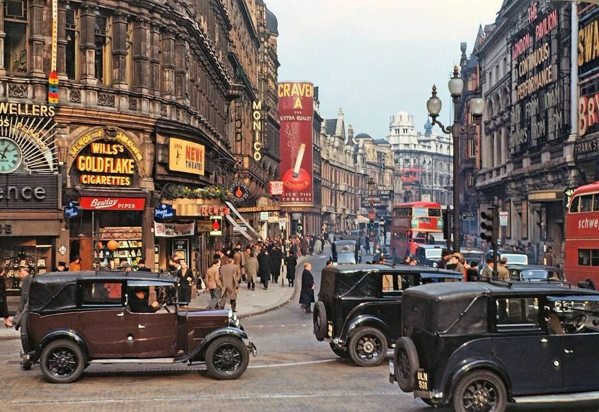 A busy city street in the mid-20th century with vintage cars, double-decker buses, people walking, and colorful shop signs advertising cigarettes, pies, and shows.