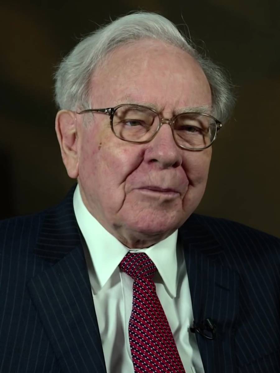An older man with white hair and glasses, wearing a dark pinstripe suit, white shirt, and red patterned tie, looks slightly off camera against a dark background.
