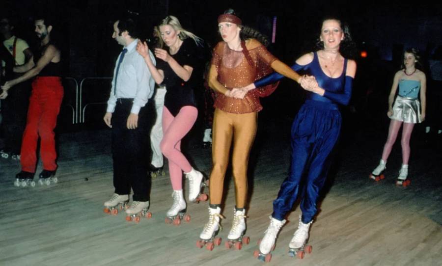 A group of people in colorful retro outfits roller skating indoors. Some hold hands while skating, and the background is dimly lit, giving a lively, vintage atmosphere.
