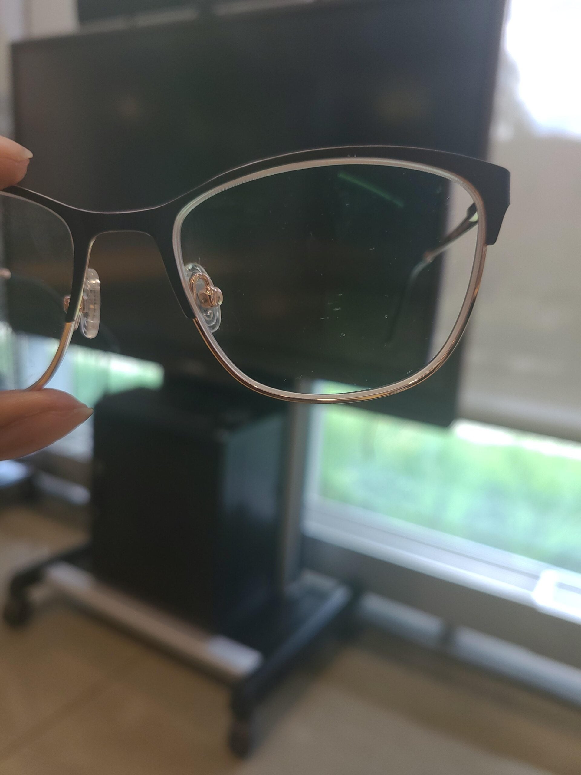 A close-up of a black-rimmed eyeglass lens held in front of a blurred TV and window, with natural light coming through and a green outdoor view visible in the background.