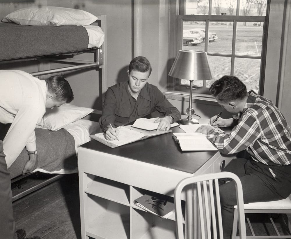 Three young men in a dorm room: one sitting at a desk writing, one reading and writing, and one making a bunk bed. Books, a lamp, and a window with cars outside are visible.