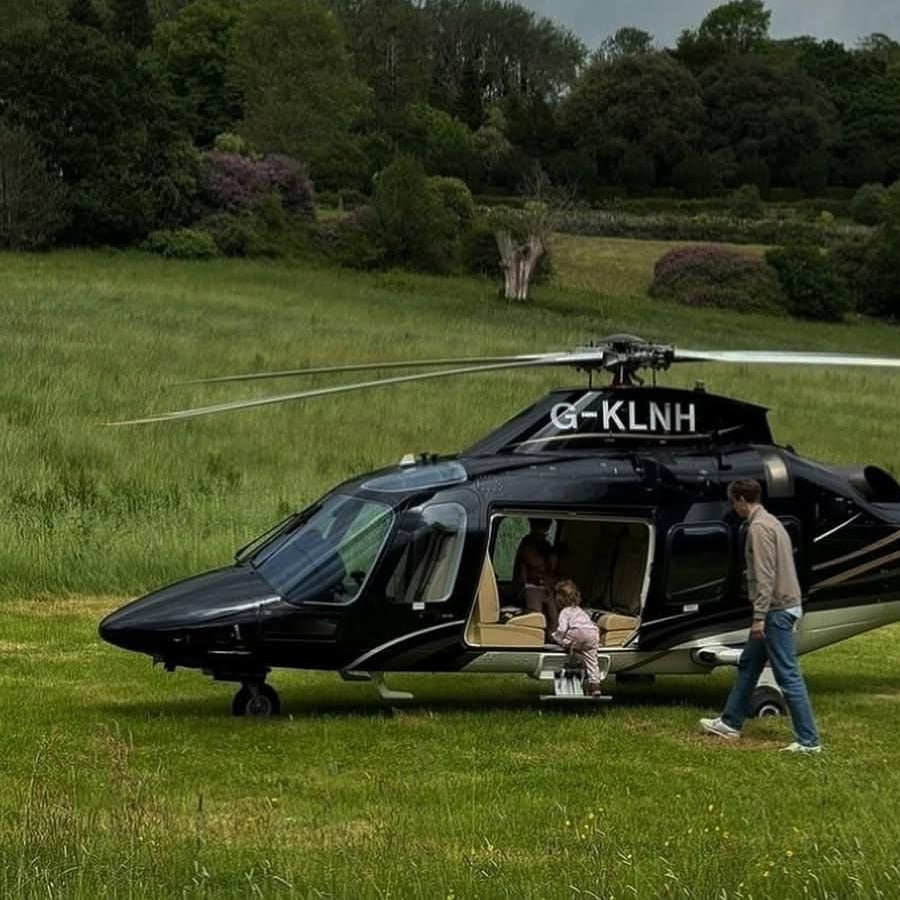 A black helicopter is parked on grass. A child sits in the doorway, and an adult stands nearby. Trees and bushes fill the background. The helicopter’s tail number, G-KLNH, is visible.