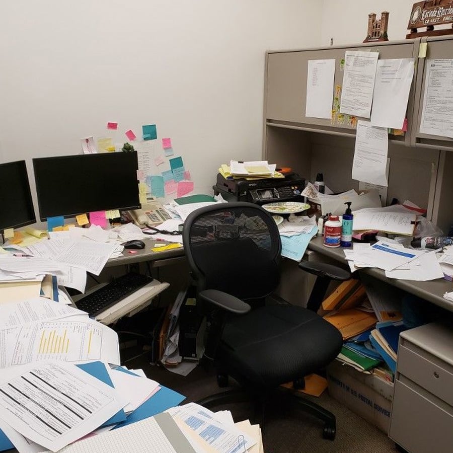 A cluttered office cubicle with messy desks covered in papers, folders, sticky notes, two monitors, office supplies, sanitizer, and a black chair. Cabinets above the desk hold more files and pinned documents.