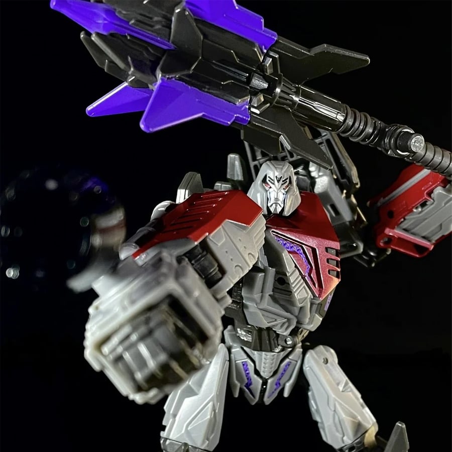 A silver and red robot action figure with a large purple weapon poses dramatically against a black background.