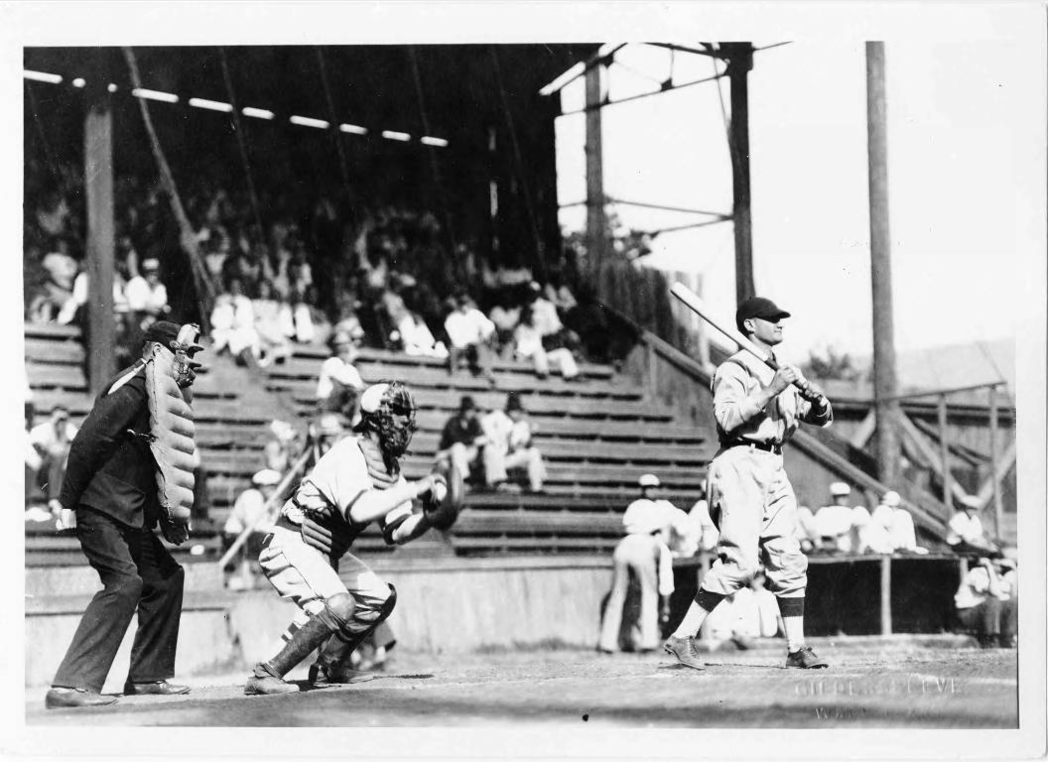 A baseball player stands at bat while the catcher and umpire watch. The background shows rows of spectators seated in a wooden grandstand.