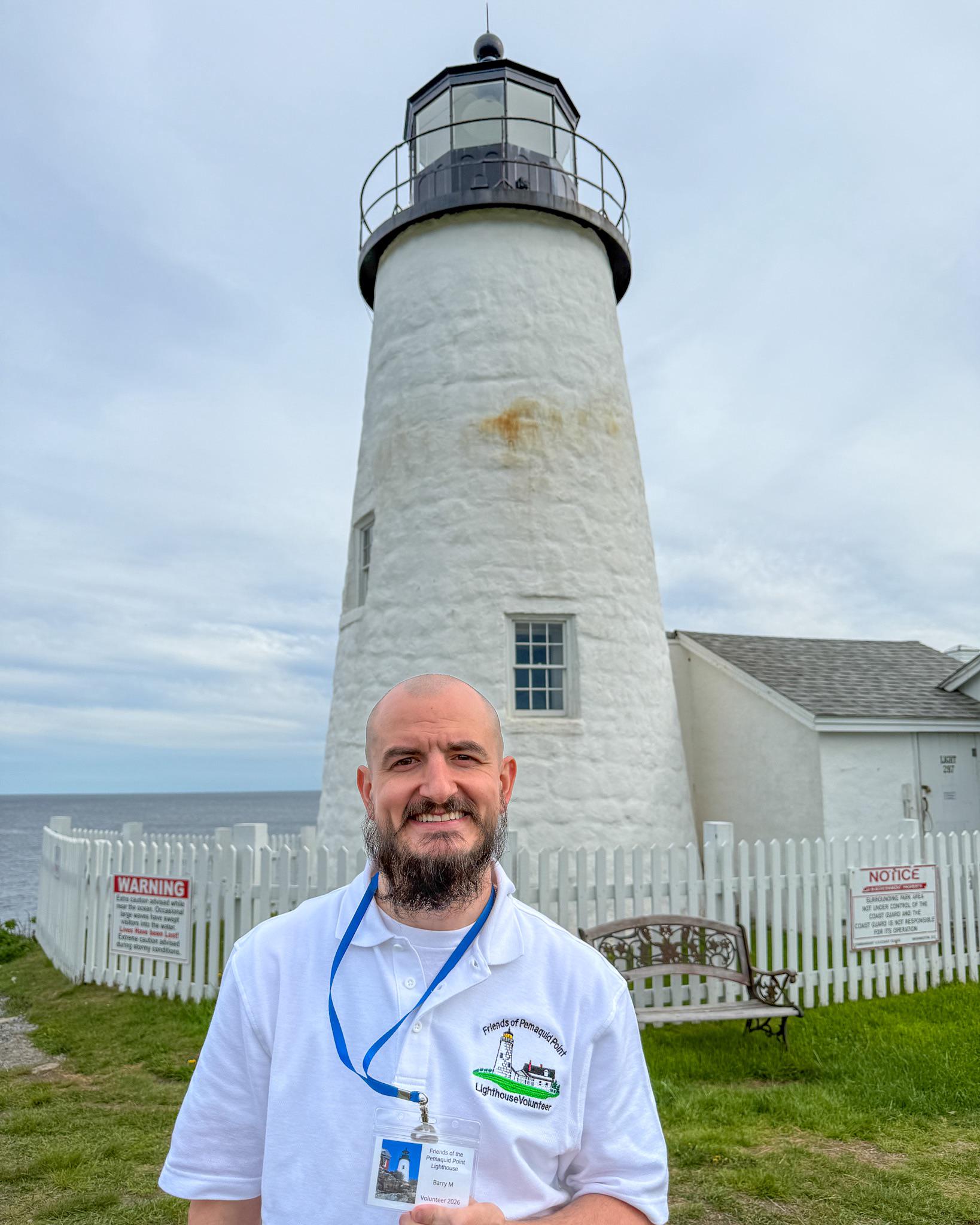 A man with a beard stands smiling in front of a white lighthouse, wearing a white polo shirt and a blue lanyard. The sky is cloudy, and warning signs are posted on a white picket fence nearby.