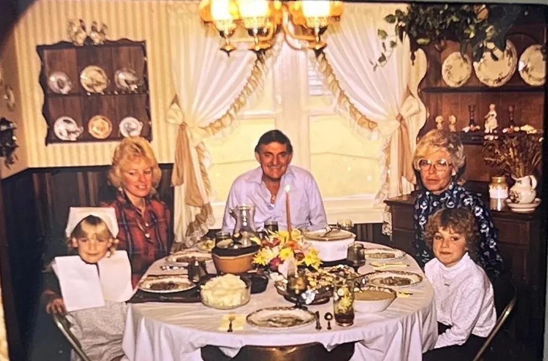 Family of five sits around a dining table set for a meal in a warmly lit room with vintage decor, flowers, and dishes. Everyone is smiling, and two children are seated at the table.