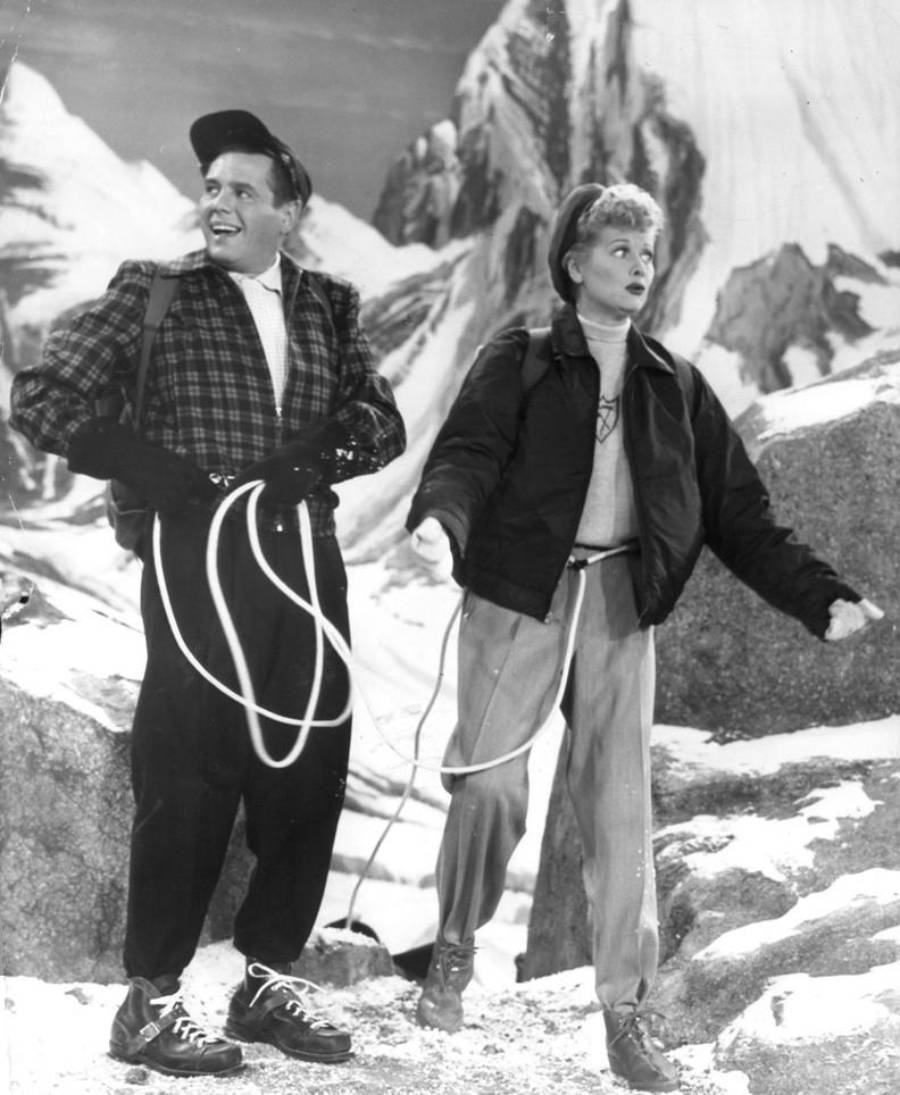 Two people dressed in hiking clothes stand on snowy rocks with ropes. The man smiles and looks left, while the woman looks confused. Tall, snow-covered mountains rise in the background.