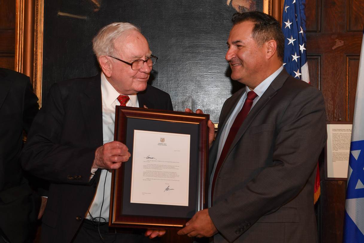 Two men in suits smile while one presents a framed document or award to the other. An American flag and another flag are visible in the background, along with a dark portrait on the wall.