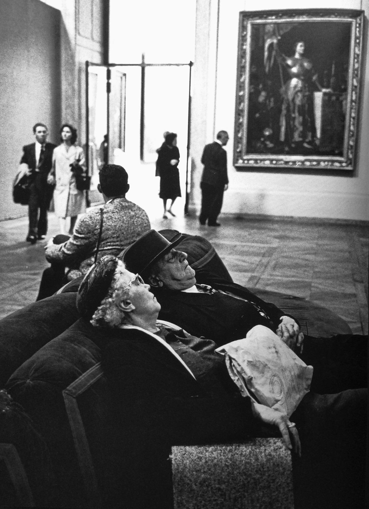 Two elderly people nap on a couch in an art museum, with other visitors walking or sitting nearby. A large framed portrait hangs on the wall in the background.